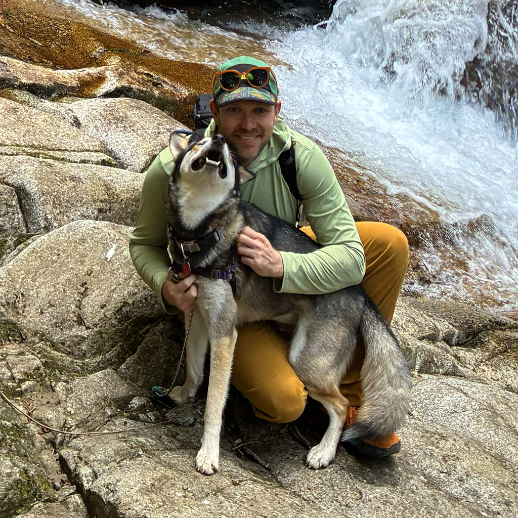 Adam crouching on granite beside a waterfall with his husky.